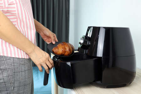 right hand of a woman is putting the homemade croissant into the tray of the black deep fryer or oil free fryer appliance which is on the wooden table in the white kitchen for the dinnerの写真素材