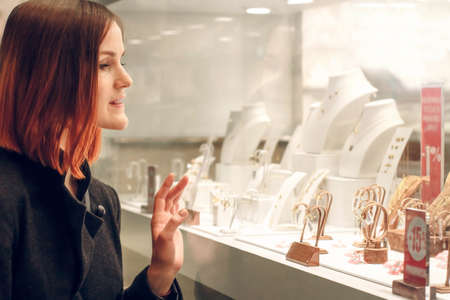 Pretty woman looking at jewelry in store window. Customer near jewelery. Dreamy red hair girl in classic style chooses silver, gold, diamonds, precious stones. Shopperの写真素材
