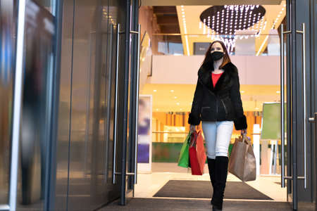 Beautiful woman in protective mask with shopping bags walks out mall. Stylish shopper girlの写真素材