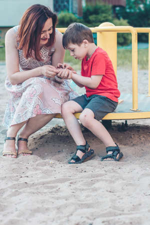 Mom and son on playground. Boy in bright red T-shirt and young woman in dress outdoors summer. Everyday life. Concept of childhood and relationship of children with parentsの写真素材