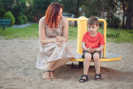 Mom and son on playground. Boy in bright red T-shirt and young woman in dress outdoors summer. Everyday life. Concept of childhood and relationship of children with parentsの写真素材