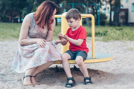 Mom and son on playground. Boy in bright red T-shirt and young woman in dress outdoors summer. Everyday life. Concept of childhood and relationship of children with parentsの写真素材