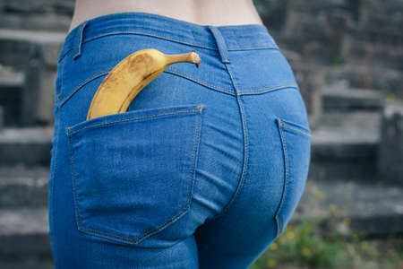 Beautiful slim ass girl in blue jeans from behind. Yellow banana in pocket booty close up. Sexy figure woman back view. Concept of sexual health, erotic, sex educationの写真素材