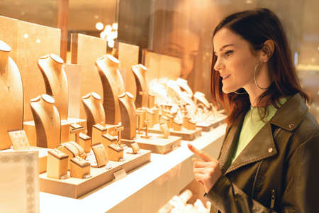 Pretty woman looking at jewelry in store window. Customer near jewelery. Dreamy sexy girl chooses silver, gold, diamonds, precious stones. Purchaserの写真素材