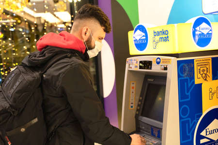 POLAND, TORUN - December 5, 2020: Man withdraws cash near ATM. Guy uses credit for money. Person in casual style wearing protective mask on his face in mall near bankomat '' Euronet ''. Debitのeditorial素材
