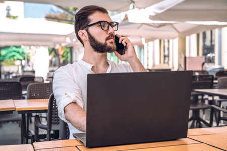 Business man with phone and laptop working in cafe. Handsome confident guy with beard in white shirt looks to side. Remote work. Freelanceの写真素材