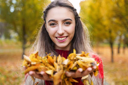 Happy smiling girl holding yellow leaves in hands. Portrait of young woman on background of park. Autumn conceptの写真素材