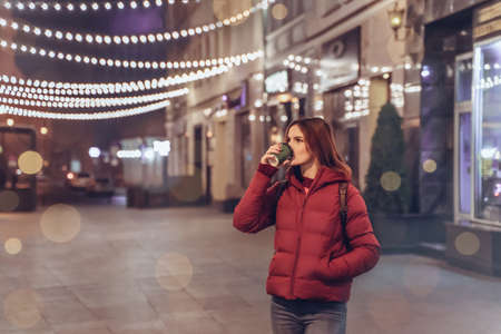 Girl in night Christmas city enjoy coffee and atmosphere. Young woman from back outdoors in winter. Lights on new years eveの写真素材