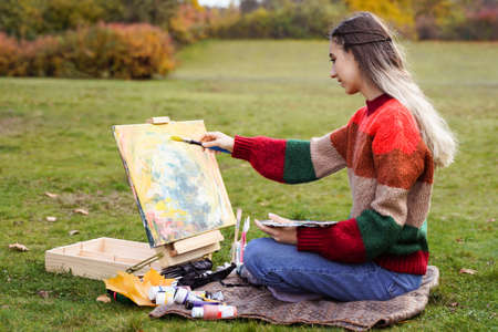 Pretty girl artist paints a picture outdoors. Woman makes sketch. Hobby creativity leisureの写真素材