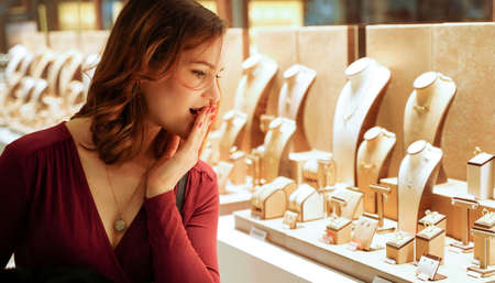 Pretty woman looking at jewelry in store window. Smiling interested customer near jewelery. Girl chooses gold, diamonds or precious stones. Buyerの写真素材
