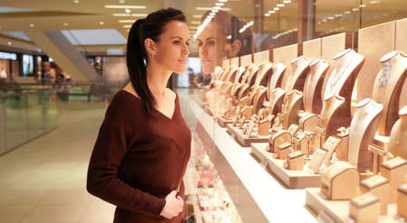 Pretty woman looking at jewelry in store window. Customer near jewelery. Dreamy long haired girl chooses silver, gold, diamonds, precious stones. Buyerの写真素材
