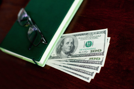 Stacks of one hundred dollars banknotes, notebook and glasses on wooden deskの写真素材