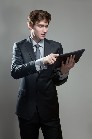 Portrait of astonished young businessman with touch screen computer, on gray backgroundの写真素材