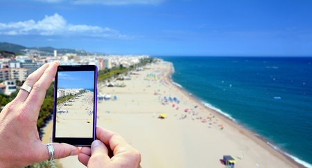 View over the mobile phone display during taking a picture of Costa Brava beach in Calella, Spain. Holding the mobile phone in hands and taking a photo. Focused on mobile phone screen.の写真素材