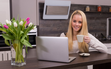 Young woman drinking coffee in kitchen with laptopの写真素材