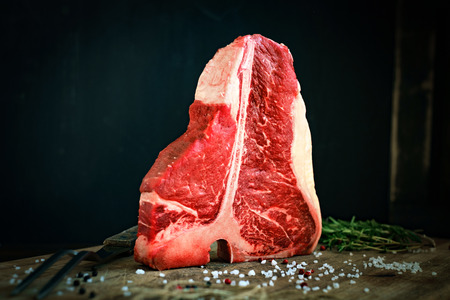 raw t bone steak is on a wooden Board on black background, next to salt, pepper and herbsの写真素材