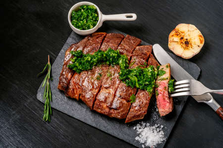 Ready to eat black Angus beef rib eye steak sliced with herbs, garlic and sauce on slate Board. Ready meal for dinner on the background of dark ebony.の写真素材