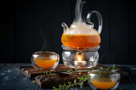 Bright tea with sea buckthorn orange in a glass teapot wooden Board on a dark background. Tea drink with vitamins and herbs.の写真素材
