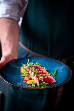 Man holds a plate of Sliced grilled tuna with green salad and tomatoes.
