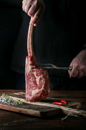 The chef's hands hold a raw Tomahawk a beef steak and a meat fork over a chopping Boardの写真素材