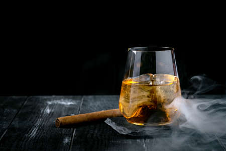 A glass of whiskey or Bourbon with ice cubes and a cigar on a black slate with smokeの写真素材