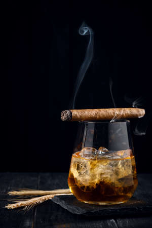 A glass of whiskey or cognac with ice cubes and a Smoking cigar lying on top, black wooden backgroundの写真素材