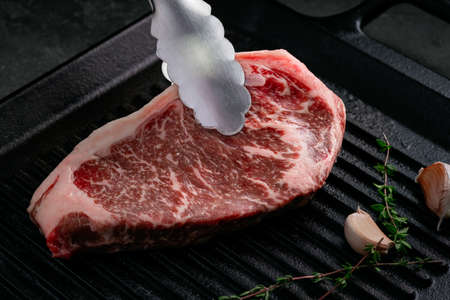 Raw Striploin steak is placed with tongs on a grill pan with garlic and thyme on a dark background, close upの写真素材