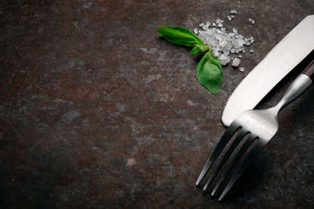 Fork and knife on a dark rusty background with salt and Basil