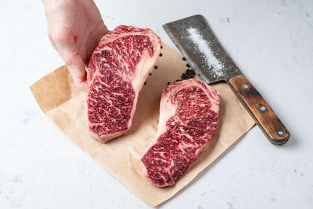 Raw marble Wagyu beef steak hold in hand, next to salt, pepper and a meat ax on a white backgroundの写真素材