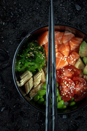 Traditional Hawaiian Poke bowl salad with vegetables and raw salmon fish, top view on black...
