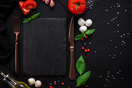 Food background for Italian cuisine. Cooking ingredients for caprese salad with fork and knife around blank slate for recipes, top view, frame, mock upの写真素材