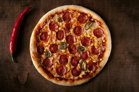 Spicy pepperoni pizza with corn and jalapeno pepper on a dark wooden background. Top view of the cooked pizza. Bannerの写真素材