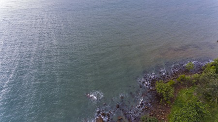 Aerial view of a rocky and green beach shore. Waves crashing on the rocks next to the lush trees....