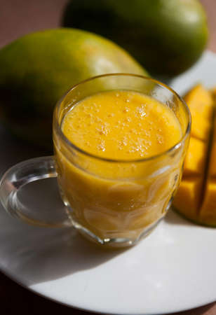The glass of fresh mango smoothie with ripe mangoes on the white plateの写真素材