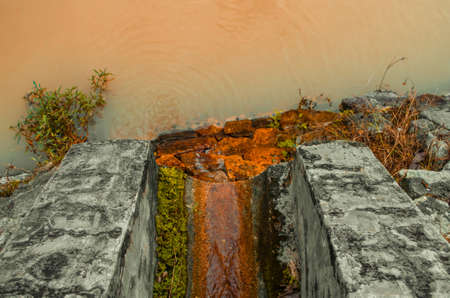 A drain with moss coverings, flowing into a river, staining the rocks below themの写真素材