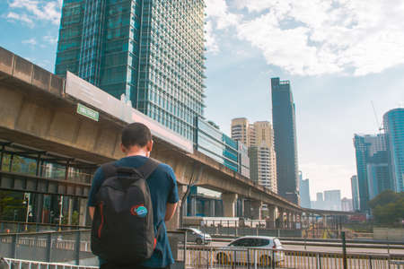 A man standing in front of a multiple-stories structure facing an overhead train track, wearing a...