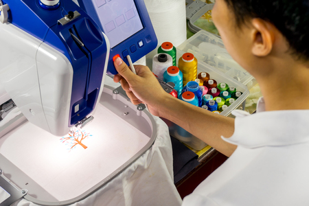 Asia man pushing red start buttom on embroidery machines next to colorful threads in plastic box and tool box on the tableの写真素材
