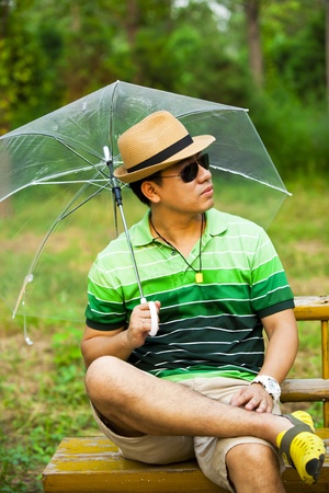 Asian young man holding an umbrella and sitting on park benchの写真素材