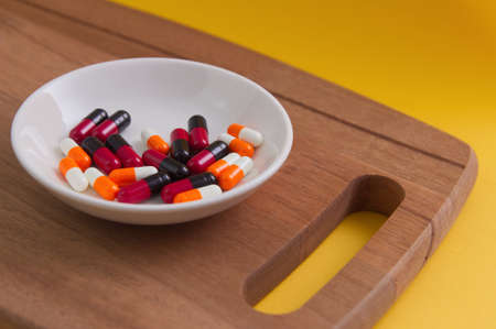 Assorted pharmaceutical medicine pills on white porcelain sauce dish plate placed on chopping board with isolated with yellow backgroundの写真素材