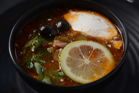 Russian traditional soup Solyanka with meat, sausages, salted cucumbers and olives. Soup in a black...