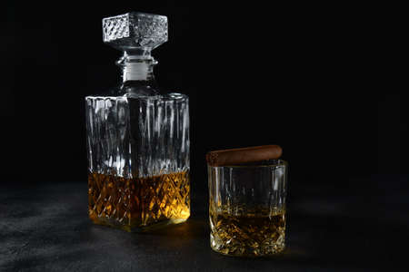 Glass of the whiskey with ice cubes and a square decanter, and cigar on a black  background.
