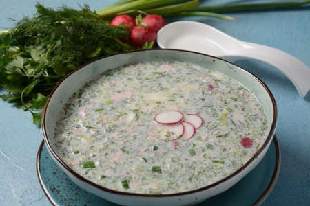 Cold soup okroshka with sausage, potato, egg, radish, cucumber, greens and kvass in a bowl, bread on wicker napkin. Traditional Russian summer cold soup with kvassの写真素材