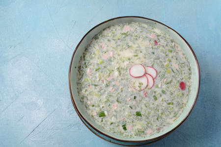 Cold soup okroshka with sausage, potato, egg, radish, cucumber, greens and kvass in a bowl, bread on wicker napkin. Traditional Russian summer cold soup with kvassの写真素材