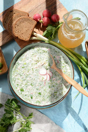 Cold soup okroshka with sausage, potato, egg, radish, cucumber, greens and kvass in a bowl, bread on wicker napkin. Traditional Russian summer cold soup with kvassの写真素材