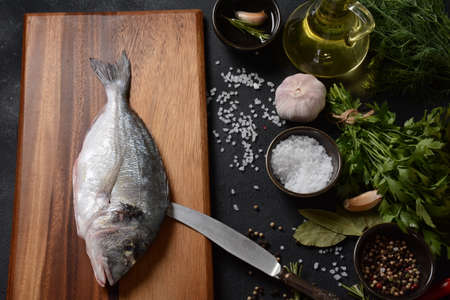 Raw fresh uncooked dorado or sea bream fish with lemon, herbs, oil, vegetables and spices on rustic wooden board over black backgroundの写真素材