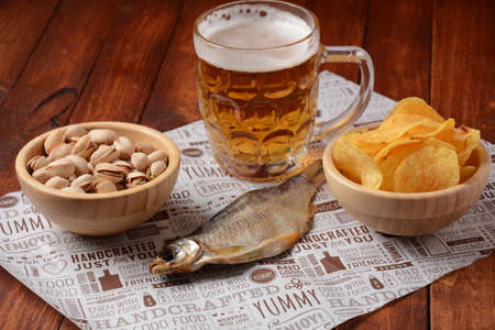 Salty dried fish roach and Beer mug with light foamed beer. Dried roach for beer on craft paper lie on a wooden table, pistachios and Potato chips in wooden bowlsの写真素材