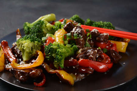 Asian teriyaki beef with red and yellow bell peppers, broccoli and sesame seeds on a plate on the table. Spicy teriyaki beef stir fry with vegetables and rice  on a dark backgroundの写真素材