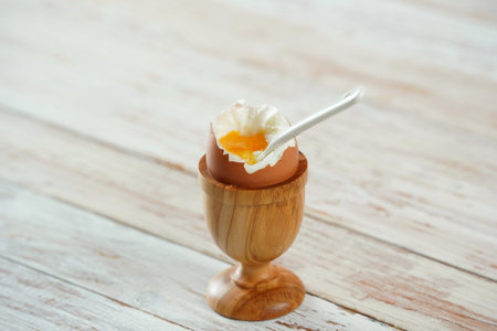 Soft-boiled egg in wooden egg cup showing runny yellow yolk with matching white ceramic spoon.の写真素材