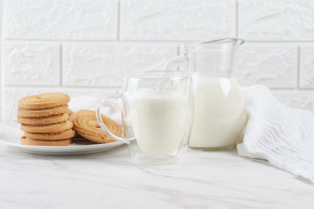 Milk in a transparent glass and jar on a marble table. Round ring shaped German spritz biscuits at backgroundの写真素材