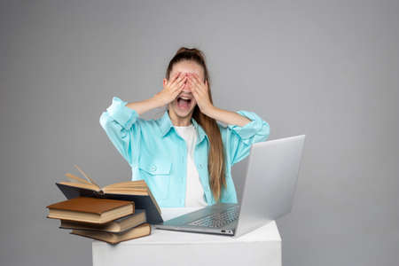 A teenage girl screams and rejoices, covering her eyes with her hands, in front of a laptop with books.の写真素材